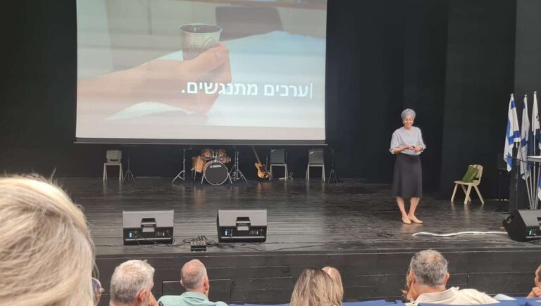 מרצה עומדת על במה באולם גדול ומעבירה הרצאה עם מצגת לקהל רחב, כחלק מפעילות הקומה הישראלית