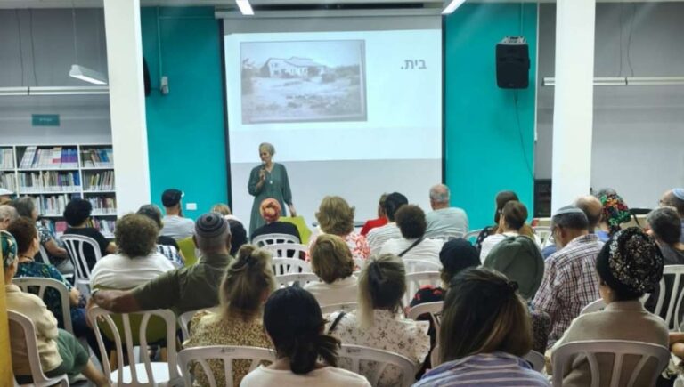 מרצה עומדת מול קהל מגוון באולם קהילתי ומעבירה הרצאה העוסקת בזהות וזיכרון במסגרת פעילות הקומה הישראלית