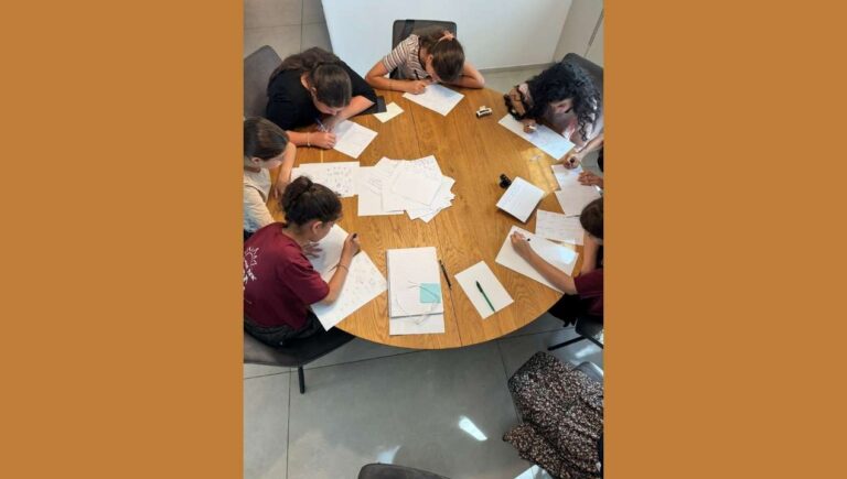 קבוצת ילדים יושבת סביב שולחן וכותבת יחד במסגרת סדנה חינוכית של הקומה הישראלית, המעודדת חשיבה ושיח ערכי